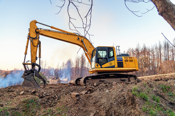 View of an excavator working in the forest on a slope. An excavator clears the slope. A fire in the forest. Smoke.