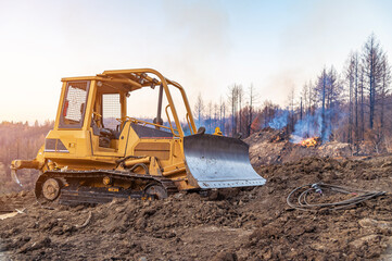 View of a bulldozer working in a forest on a slope. The bulldozer clears the slope. A fire in the forest. Smoke. - Powered by Adobe