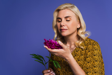 blonde woman in blouse smelling purple flower with green leaves isolated on violet.