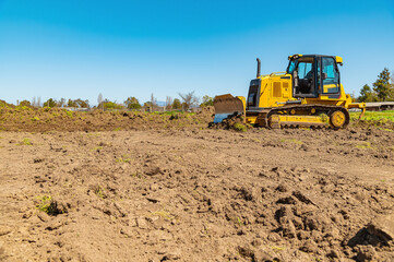 Sidet view of a yellow bulldozer on a plot of land. A bulldozer clears the area. Blue sky background. - obrazy, fototapety, plakaty