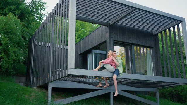 Young couple sitting and cuddling in hammock terrace in their new home in tiny house in woods, sustainable living concept.