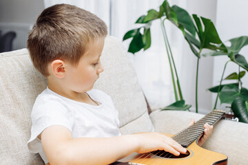 Adorable little boy learning to play acoustic guitar at home. Hobbies and entertainment for...