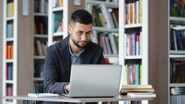 Handsome young man with eyeglasses sitting in library and browsing internet on laptop, looking stressed and unhappy. Distance learning. Remote work. Concept of technology