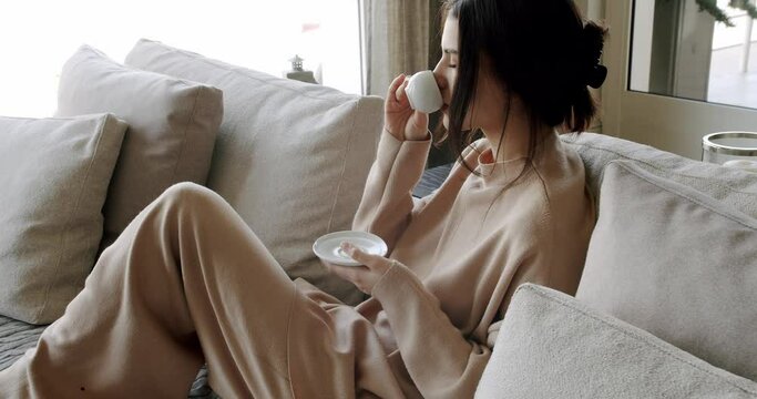 A beautiful Young woman is drinking coffee and tea in the living room at home, healthy calm lady relaxing on comfortable sofa feel stress free at home lounge alone. Woman rests at home on luxury sofa.