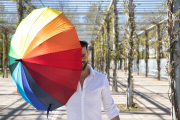 Young, handsome gay man with blue eyes, perfect smile and wearing a white shirt, covering his face with a rainbow-colored umbrella. Concept gay pride, homosexual, lgtbi, pride day.