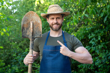 Happy farmer man in farmers hat and apron pointing finger at garden spade natural outdoors