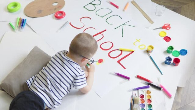 little smart preschooler boy lying on floor drawing back to school phrase colorful pens crayons on white paper. creative activity kindergarten indoors. art class at home education family concept.