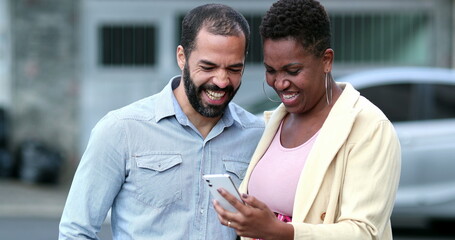Interracial couple laughing in street holding cellphone. Candid mixed couple relationship looking at smartphone. Real life laugh and smile