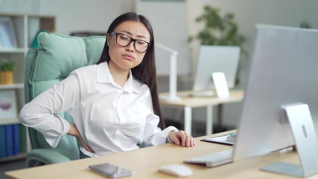 young asian woman affected back ache at the workplace. Female backpain sitting for a long time at the computer desk in the office. Indoor. An employee entrepreneur suffers from pain tension. 