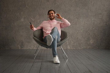 Handsome man listening to music while sitting in comfortable armchair against gray wall.