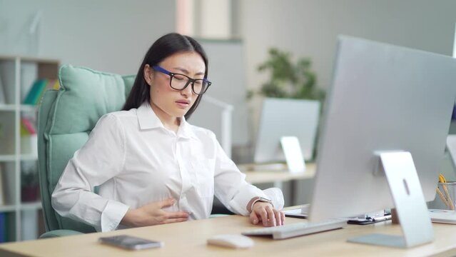 Young Asian woman worker, employee entrepreneur with stomach pain, poisoning, Premenstrual syndrome, heaviness in abdominal Suffering sitting at workplace at work in office at computer desk. Indoor