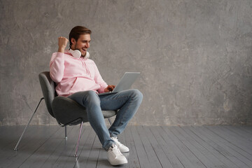 Joyful Male Using Laptop And Shaking Fists Celebrating Great News And Luck Sitting In Chair Over Gray Wall Background