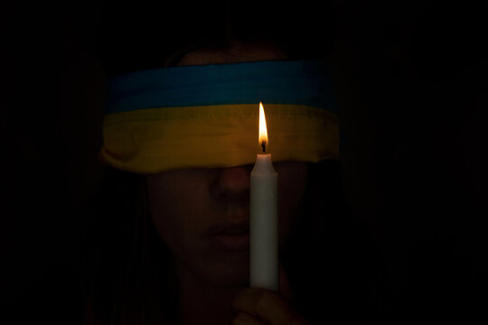 A girl blindfolded with the flag of Ukraine and a candle in her hands in the dark, the flag of Ukraine in front of the girl, war and fear of people 2022