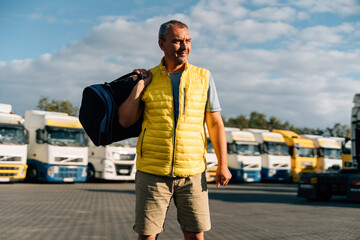 Portrait of caucasian mature man with bag on some-truck vehicles parking background. Truck driver worker 