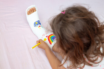 A unrecognizable little girl with a broken leg at home on the bed draws with felt-tip pens on an...