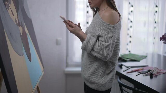 Unrecognizable young woman standing at easel with picture surfing social media on smartphone. Side view slim Caucasian artist searching inspiration online. Art and modern technologies