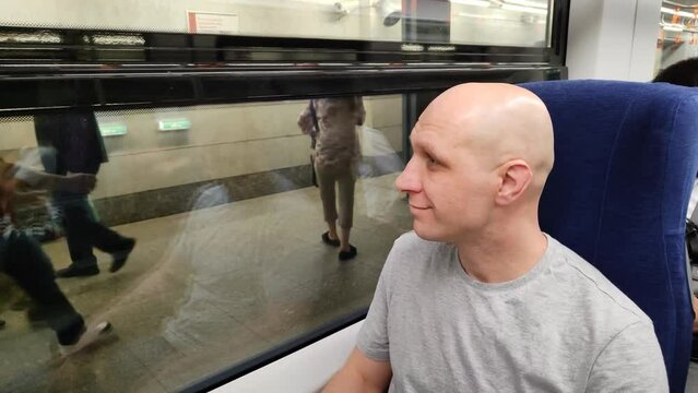 Young man sitting in train, looking out window and waving to people seeing off him. Bald guy in subway or train car. Moscow Central Ring (MCC), Lastochka train. Train on platform. Modern transport