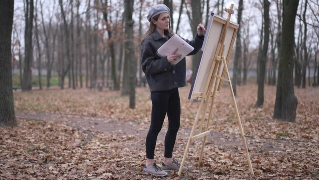 Wide shot focused talented young woman walking to easel in autumn park making strokes on canvas with brush. Concentrated confident Caucasian painter creating picture outdoors on overcast day