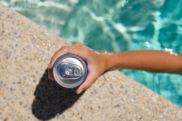 Can of cold drink in hand at the edge of the hotel pool. Top view, selective focus on the bank.