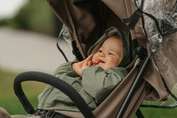 A photo from the side of a female toddler who is lying in the stroller on a cloudy day. A young girl in the raincoat is in her baby carriage in a park at noon.