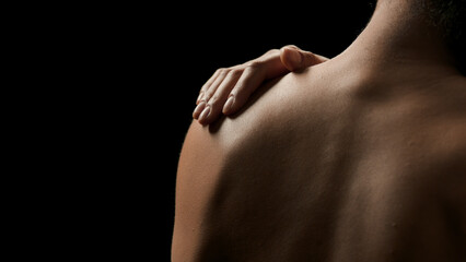 Fototapeta premium Horizontal close-up shot of young good-looking slim African American woman touches her bare shoulder on black background | Body care concept