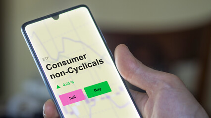 Un investisseur analyse le fonds ETF de consommation non cyclique sur un écran. Un téléphone affiche les prix des biens de consommation non cycliques, texte en anglais.