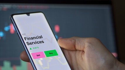 Un investisseur analyse le fonds ETF des services financiers sur un écran. Un téléphone affiche les prix des services financiers, texte en anglais.