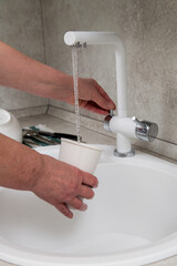 The hands of an adult woman pour water from the tap into a glass in the kitchen