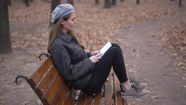 Inspired talented young woman drawing sketch sitting on bench walking away in autumn park. Slim beautiful Caucasian artist creating idea outdoors on overcast day. Inspiration and lifestyle