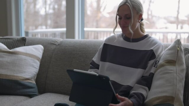 woman walking to the couch to lay down and scroll through tablet while drinking coffee in a nice house. followed with camera from the side closer up.