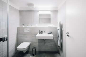 A small modern bathroom with a large mirror, square sink, toilet and glass shower enclosure. There are also some additional items as towel, candles, soap dispenser, waste bin, etc.