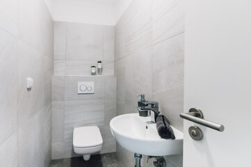Small modern toilet with sink, towel and soap dispenser. On the surrounding ones, we can find a switch, a flush button and the part of the door with a handle and lock visible to the right.