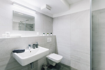 Small simple bathroom with mirror.  Bathroom has square sink and shower with glass door and toilet with seat closed. There are also some additional items as bin, dispenser, towel, etc.