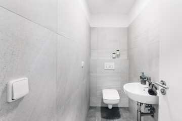 Small modern toilet with sink, towel and soap dispenser. On the surrounding ones, we can find a switch, a flush button and the part of the door with a handle and lock visible to the right.