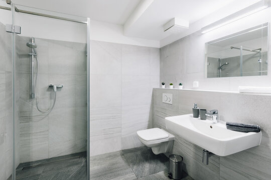 Small simple bathroom with mirror.  Bathroom has square sink, large and spacious shower with glass door and toilet with seat closed. There are also some additional items as bin, dispenser, towel, etc.