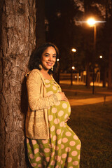 pregnant latin mom leaning on a tree in a park at night looking at camera