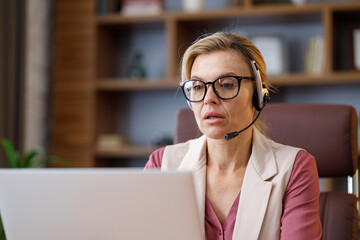Smiling businesswoman wearing headset talking to laptop web camera sitting in office. Recording vlog, online video conference, video call, online education. Job interview, customer support