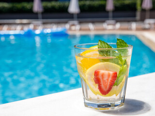 Citrus mint leaves infused water, cocktail, lemonade in glass near pool. Summer iced cold drink with lemon, lime, orange, strawberry, mint on blue water white marble background, copy space, side view
