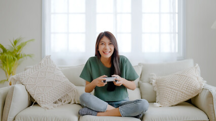Beautiful young asian woman sitting in living room sofa holding joystick remote control playing...
