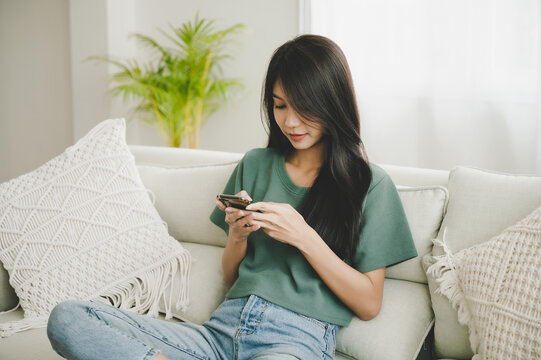 Happy young asian woman relaxing at home she is sitting on sofa and using mobile smartphone