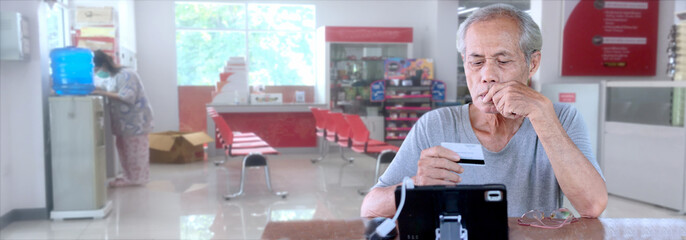 Asian elderly man sitting in front of a tablet holding a credit card in a shop