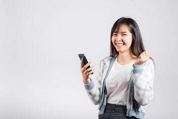 Woman excited holding mobile phone funky successful raise fists say yes! studio shot isolated white background, happy young female smiling surprised make winner gesture on smartphone