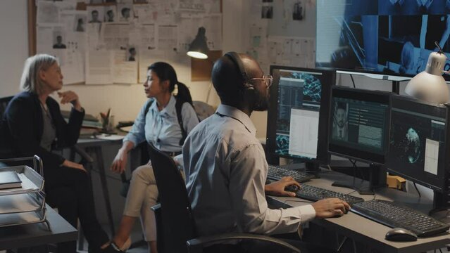 Medium long of young Black man using computer, searching federal criminal database, two female detectives sitting and talking on background in office