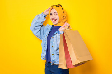 Happy young Asian woman in jeans jacket holding shopping bags and touching sunglasses, looking at camera isolated over yellow background
