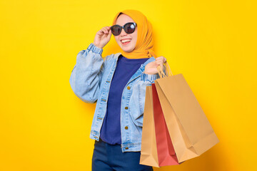 Happy young Asian woman in jeans jacket holding shopping bags and touching sunglasses, looking at camera isolated over yellow background