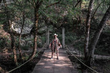 Mexican young man wearing a poncho and a hat is standing with a duel pose on a wooden bridge