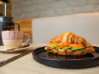 croissant with red fish and cucumber on a plate and a cup of coffee on a wooden table