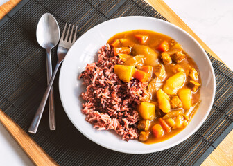 Japanese curry with rice berry in round white plate, healthy food with natural light, Japanese curry and rice with spoon and fork black wooden placemat, view from above.