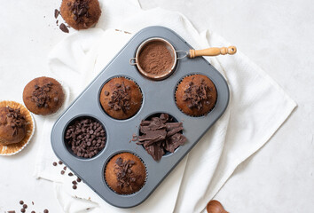 Close up top view of chocolate muffins flat lay in baking tray with slides of chocolate, chocolate chip and cocoa powder on white cutting board and white cloth