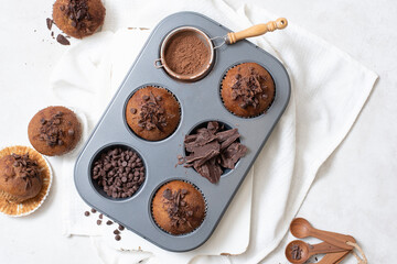 Close up top view of chocolate muffins flat lay in baking tray with slides of chocolate, chocolate chip and cocoa powder on white cutting board and white cloth